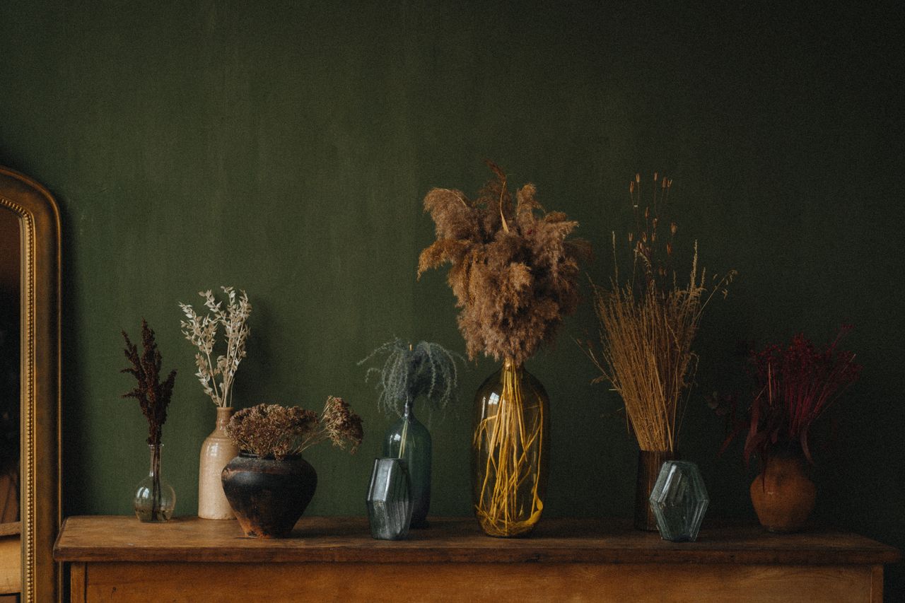 Dried garden stems in collected vases against an earthy green wall